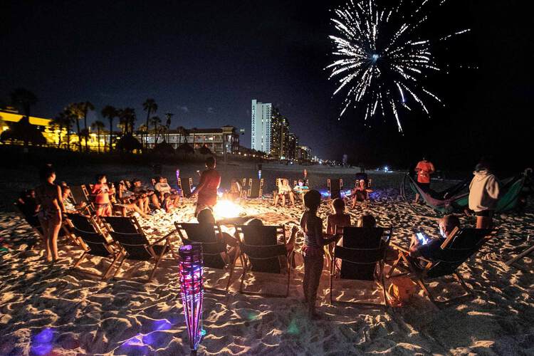 Group seated around bonfire on beach at night with fireworks in sky
