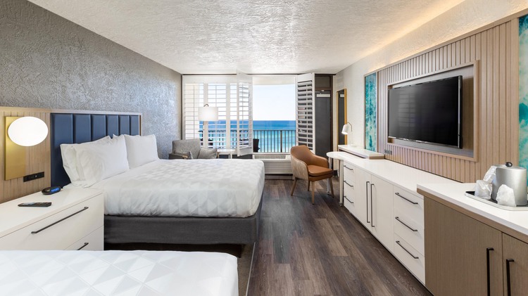 Two queen beds with mounted TV, work desk, chair and view of ocean from balcony